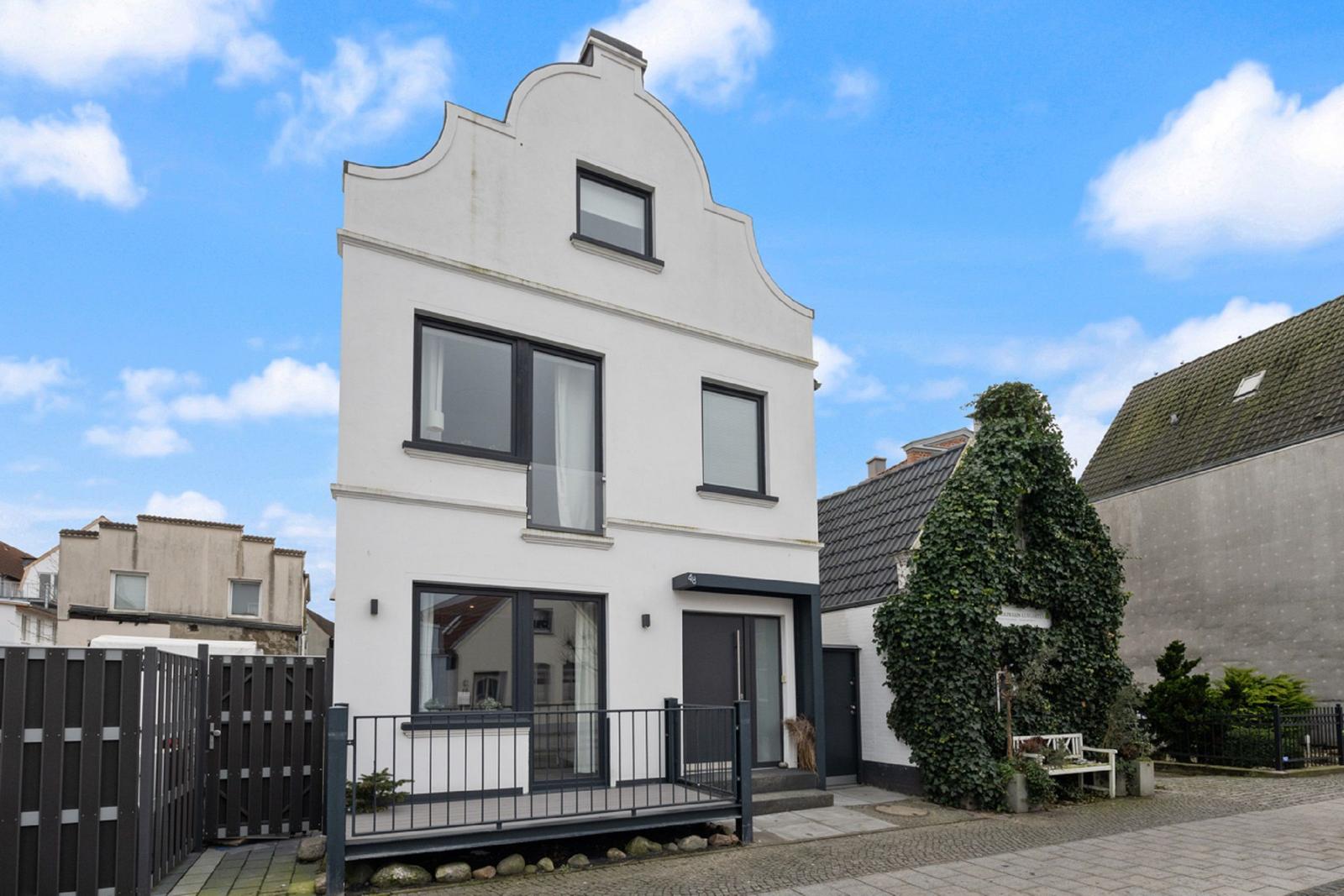 Weiße Hausfassade mit schwarzem Fensterrahmen und Vordereingang. Gartenbereich mit Bänken und Pflanzen.