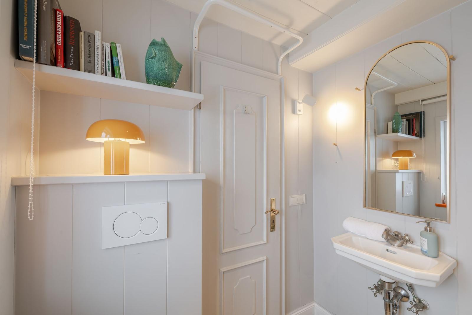 Badezimmer mit Spiegel, Waschbecken und Toilette. Regal mit Büchern und Lampe.