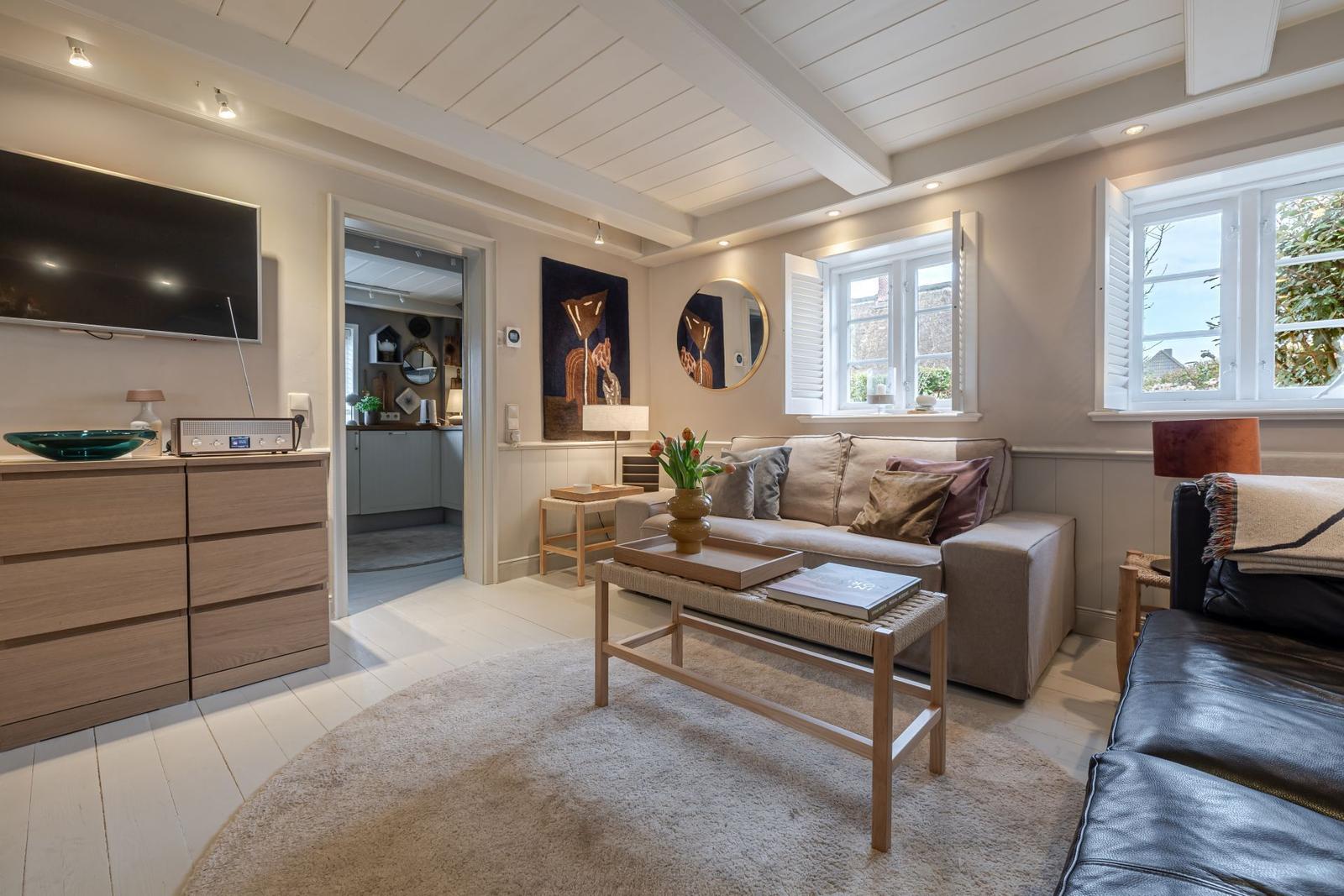 Wohnzimmer mit Sofa, Couchtisch und TV. Durchgang zur Küche. Fenster mit Jalousien.