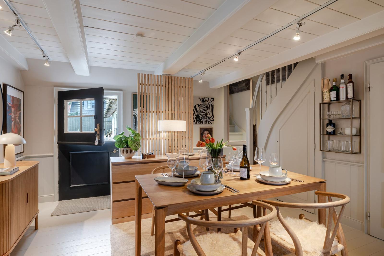 Esszimmer mit Holztisch, Stühlen und Weinflaschen. Helle Wände, Deckenbalken und Treppe sichtbar.