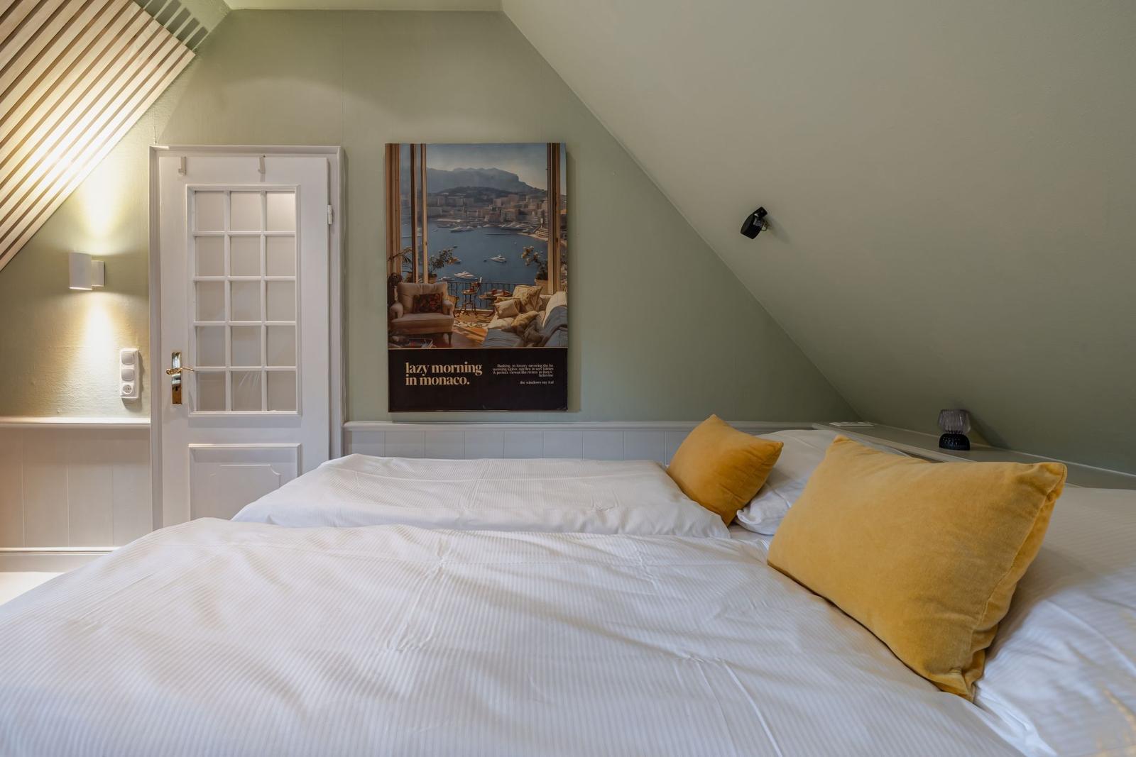 Schlafzimmer mit Dachschrägen, Bett mit gelben Kissen und Wandbild.