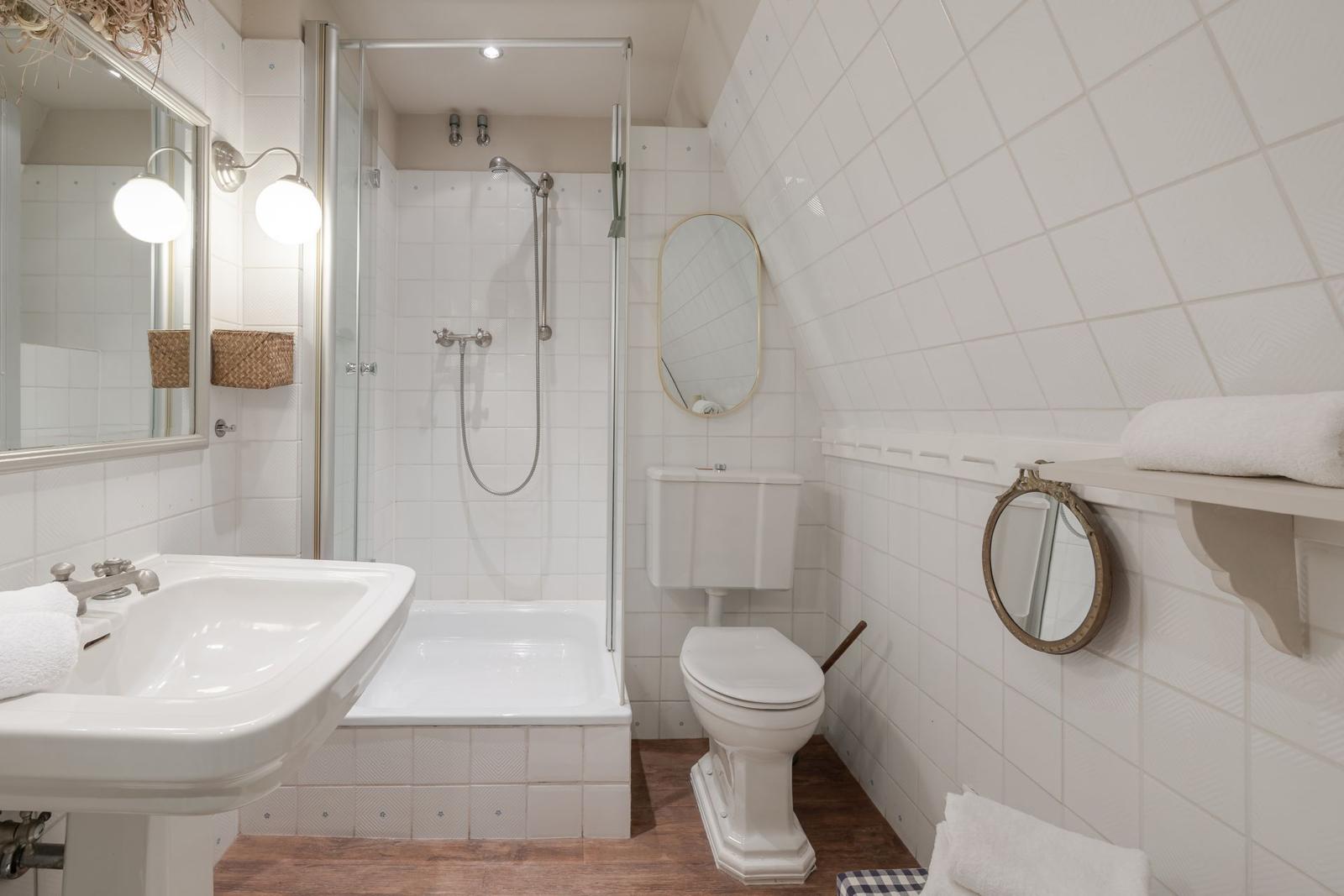 Badezimmer mit Dusche, Waschbecken und Toilette. Weiße Fliesen und Holzboden.