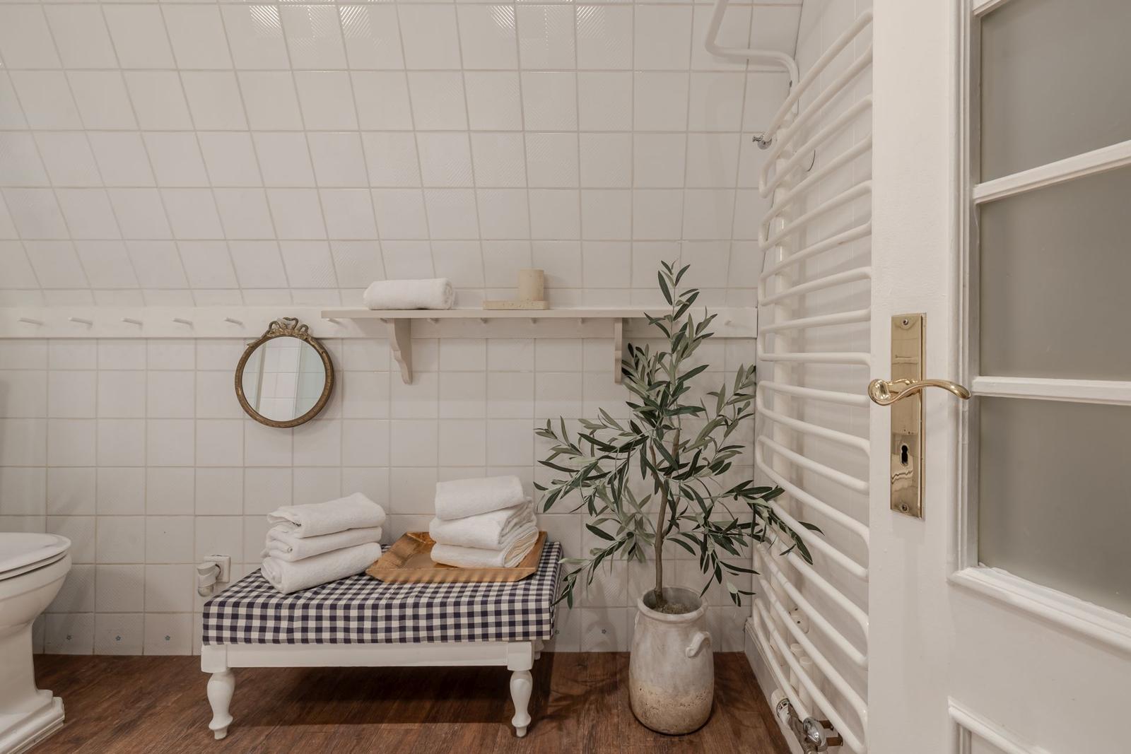 Badezimmer mit weißen Fliesen, Holzboden, Toilette, Spiegel und Pflanze.