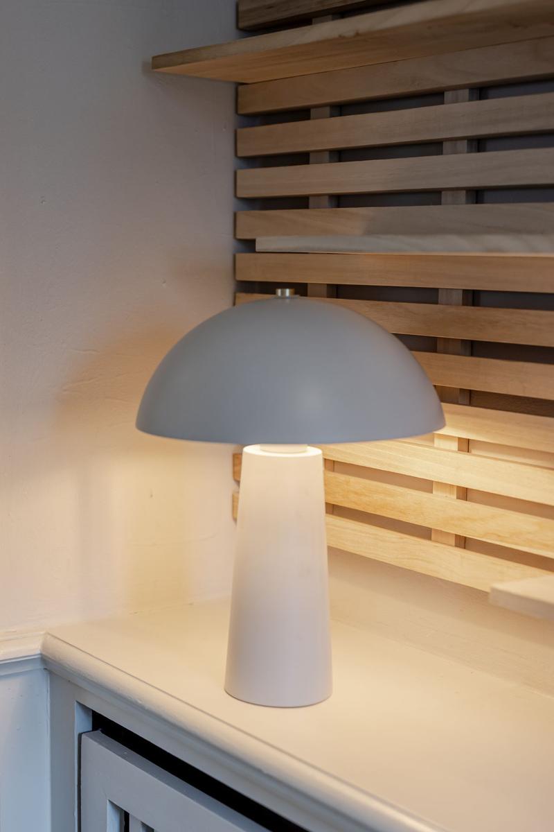 Eine Tischlampe mit grauem Schirm steht auf einer weißen Oberfläche vor einer Holzleistenwand.