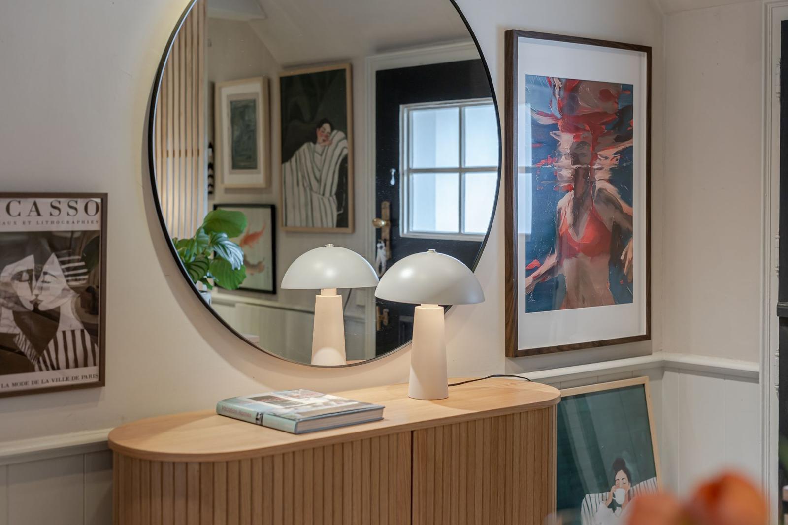 Rundes Spiegelbild, zwei weiße Lampen und ein Holzschrank mit Bildern an der Wand.