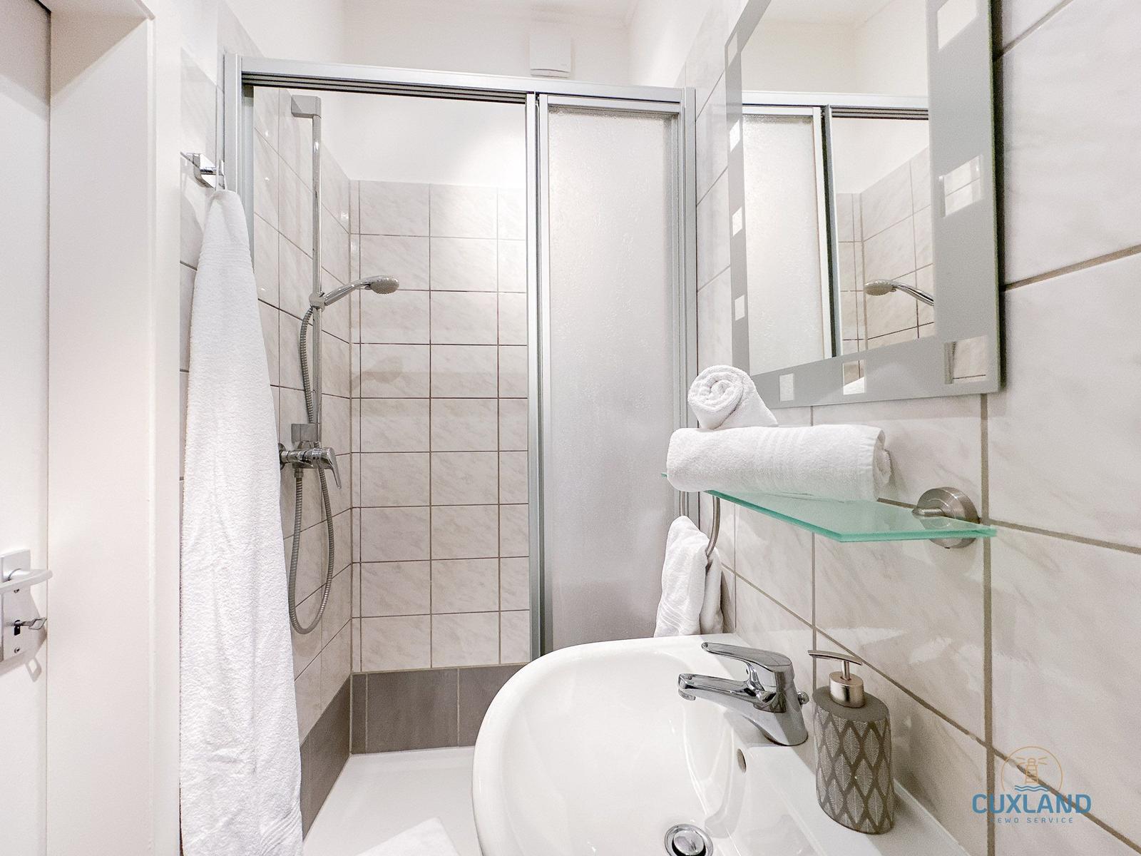 Badezimmer mit Dusche, Waschbecken und Spiegel. Handtücher und Seife sind vorhanden.