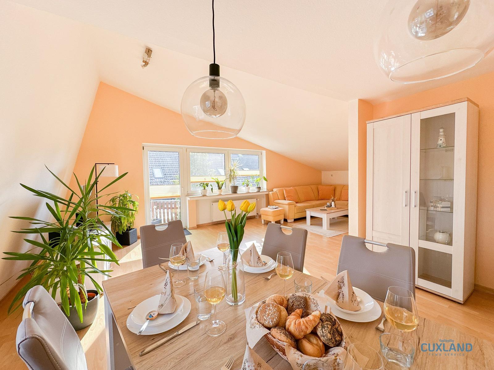 Esszimmer mit Tisch, Stühlen und Blumen. Im Hintergrund Wohnzimmer mit Sofa und Fenster.