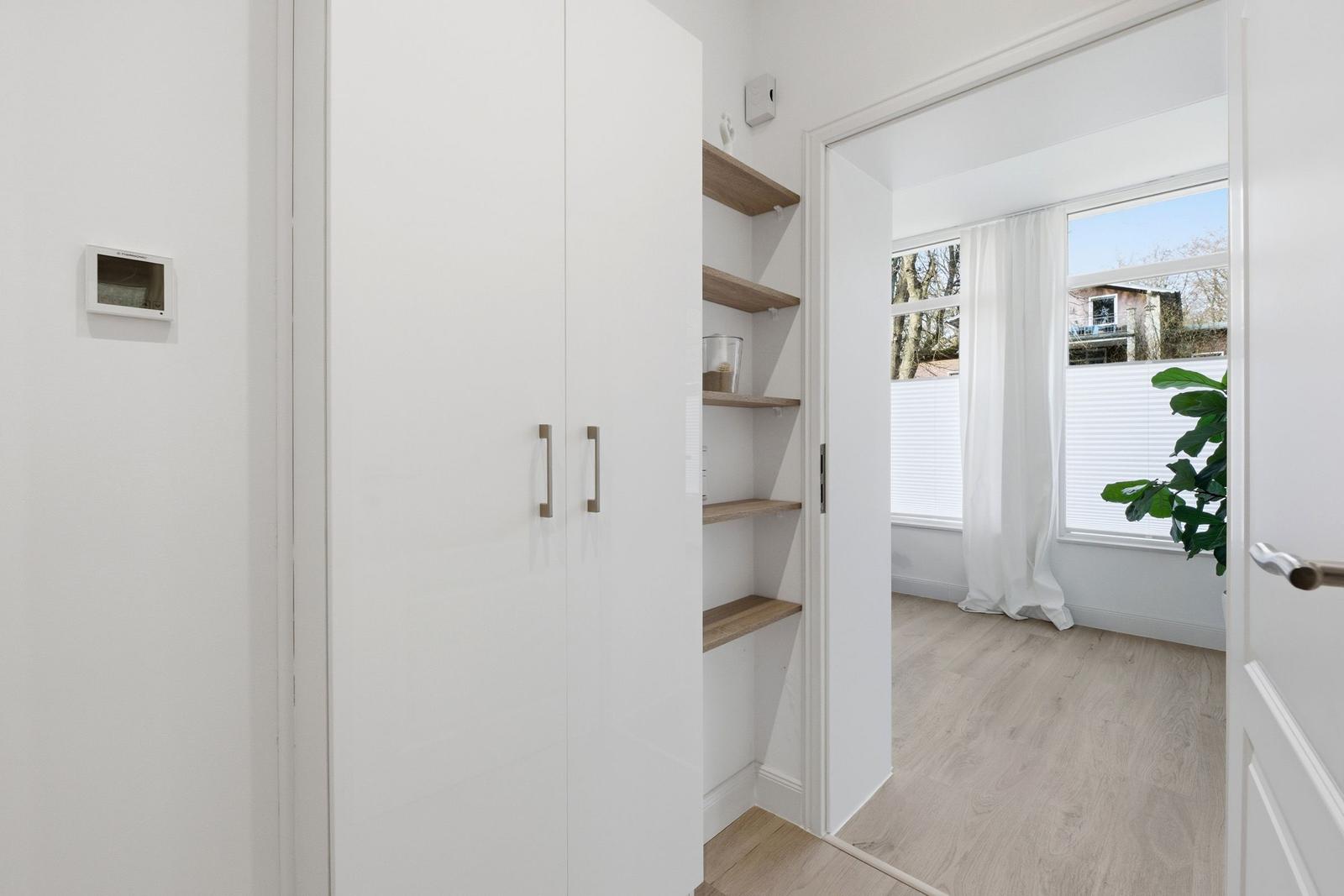 Schlafzimmer mit weißen Schränken und Holzregalen. Fenster mit Vorhängen und Pflanze.