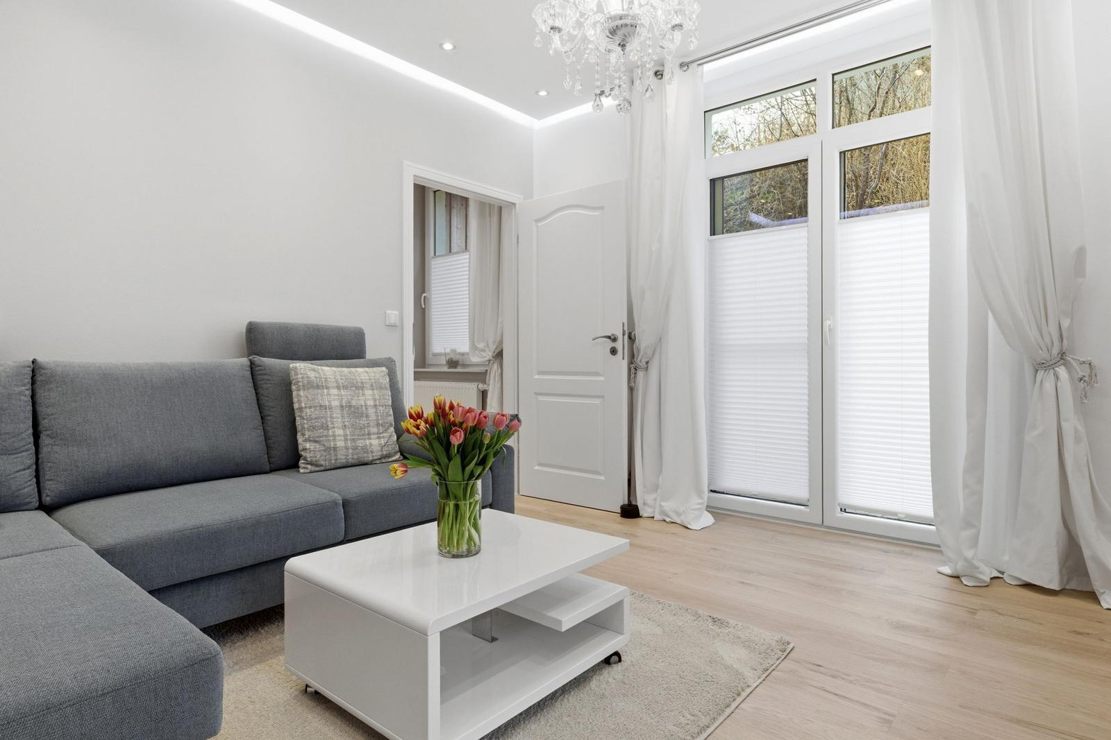 Wohnzimmer mit grauem Sofa, weißem Couchtisch und Blumen. Große Fenster mit weißen Vorhängen.