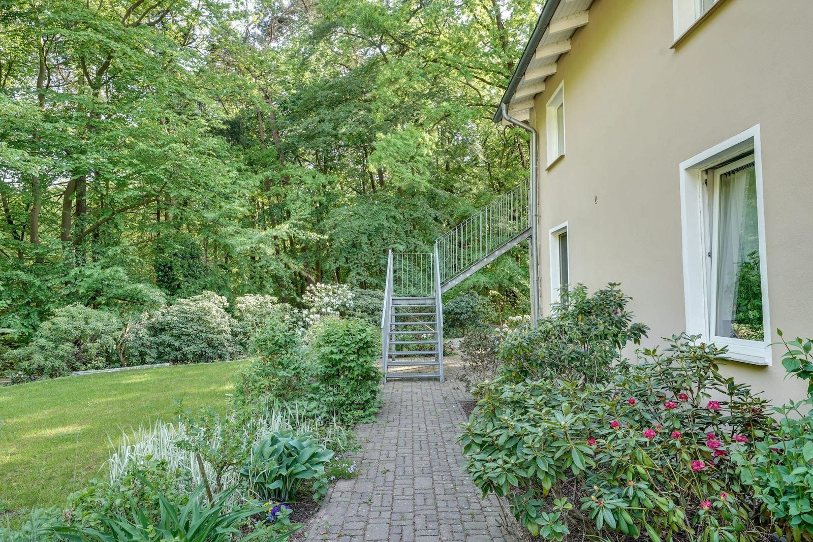 Haus mit Garten, Steg und Blumenbeete. Wald im Hintergrund.