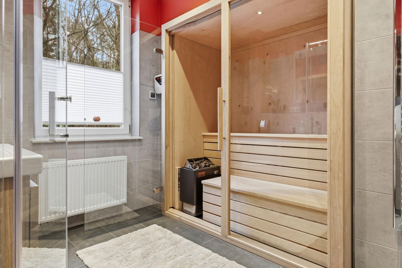 Sauna mit Holzbänken und Glaswand. Fenster mit Jalousie. Fliesenboden und Teppich.
