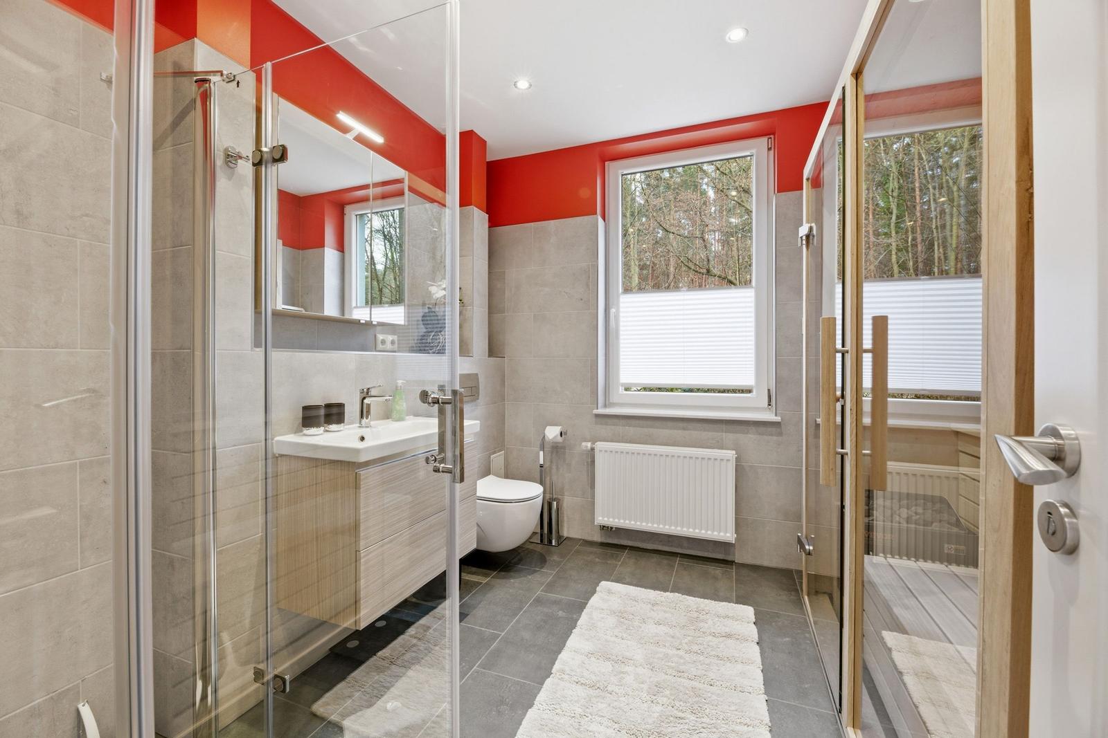 Badezimmer mit Dusche, Waschbecken und Toilette. Fenster mit Blick auf Wald.