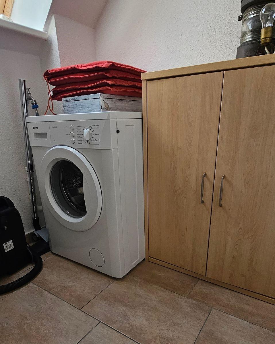 Waschmaschine neben einem Holzschrank mit rotem Kissen darauf.