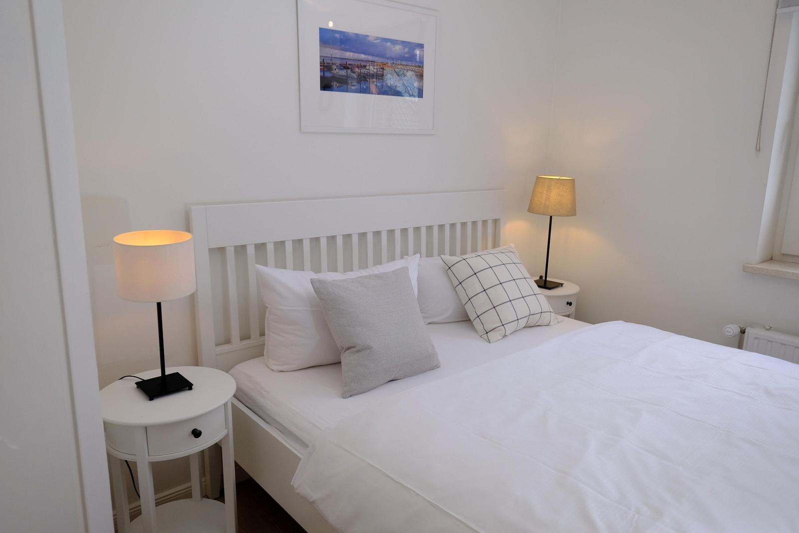 Schlafzimmer mit weißem Bett, zwei Nachttischen und Lampe. Wandbild zeigt Hafen.