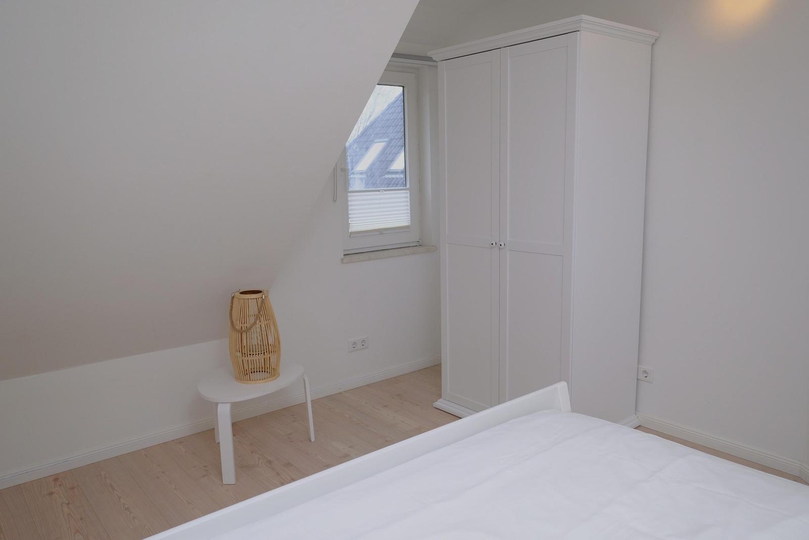 Schlafzimmer mit weißem Bett, Kleiderschrank und Fenster mit Blick auf Dach.