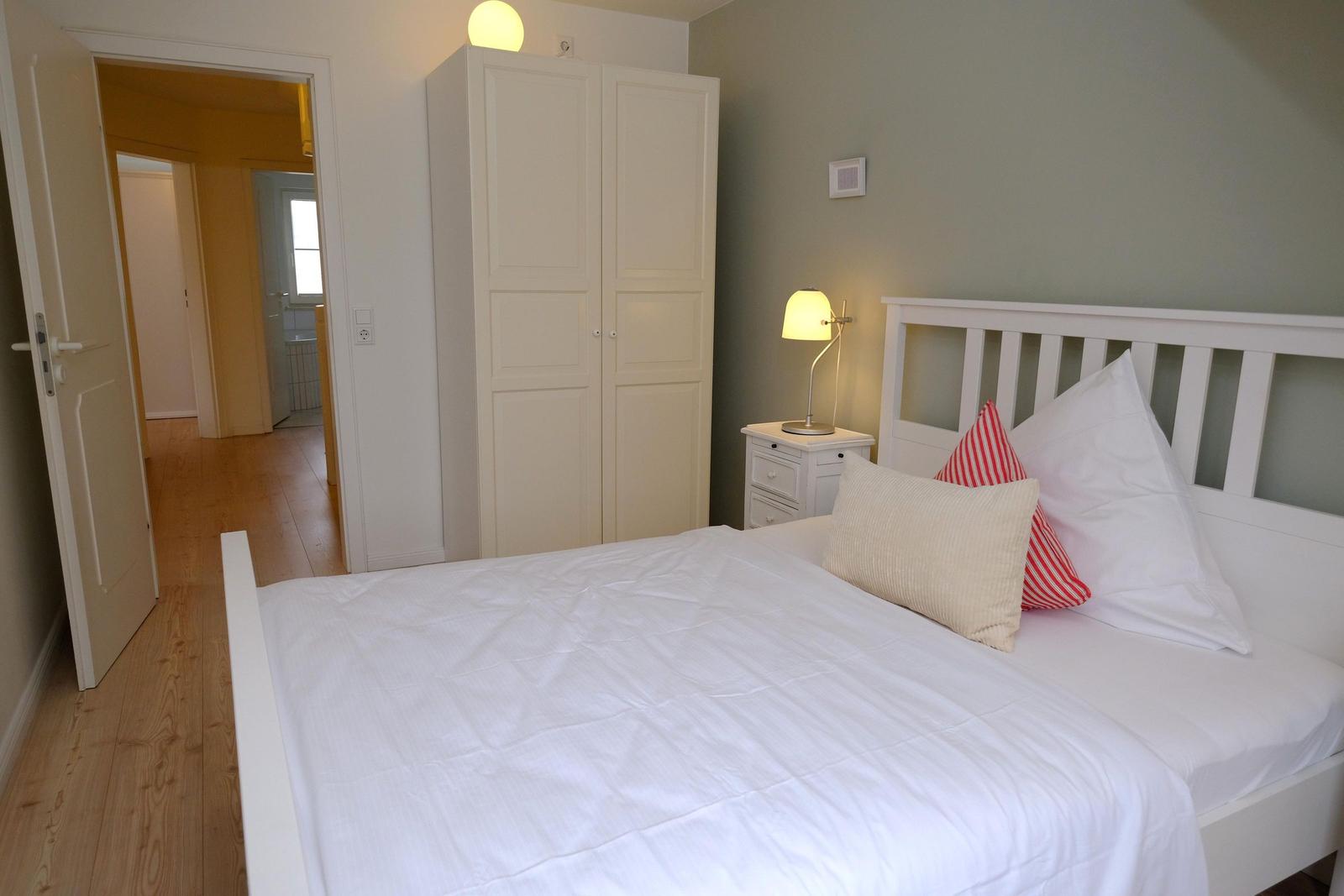 Schlafzimmer mit weißem Bett, Nachttisch und Kleiderschrank. Holzboden und Tür zum Flur.