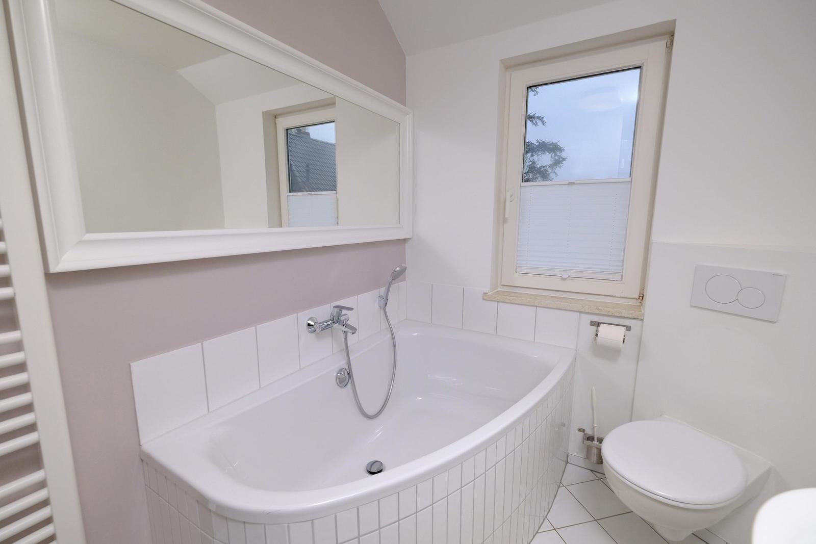 Badezimmer mit Badewanne, Spiegel und Toilette. Fenster mit Jalousie.