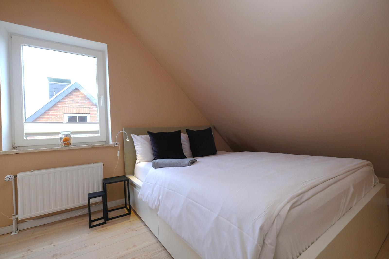 Schlafzimmer mit Dachschrägen, weißem Bett und Fenster mit Blick auf Dach.