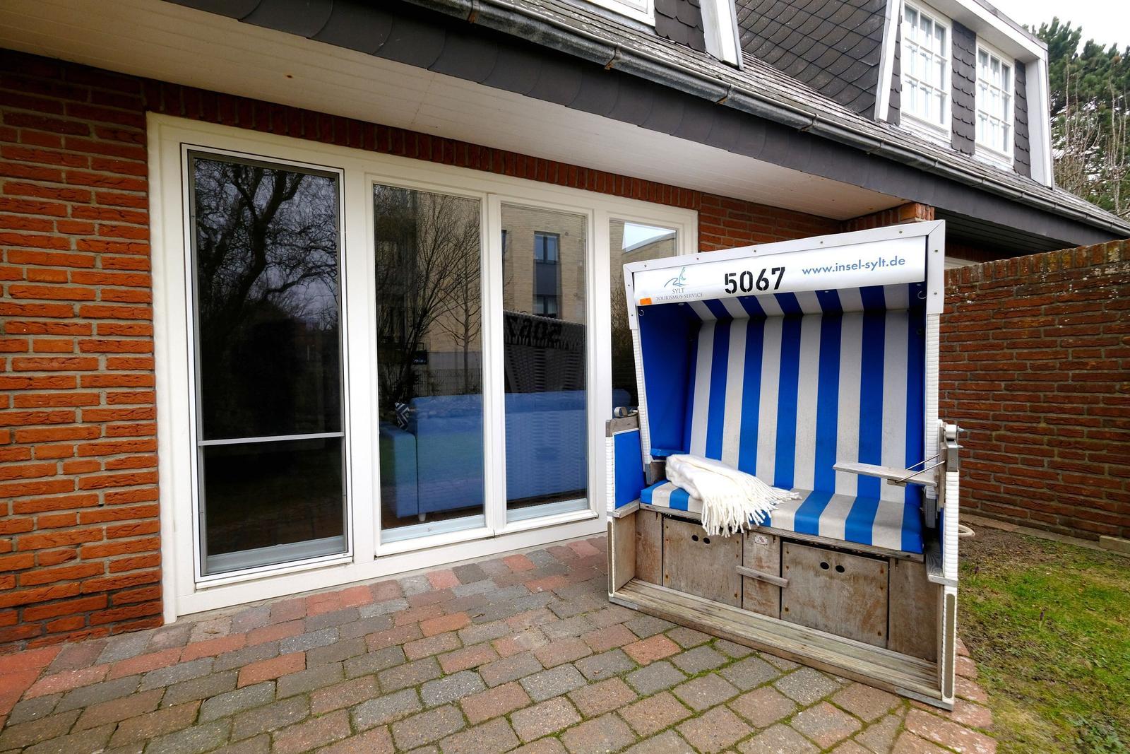 Blau-weiße Strandkorb mit Nummer 5067 vor Haus mit Fenstern und Ziegelmauer.