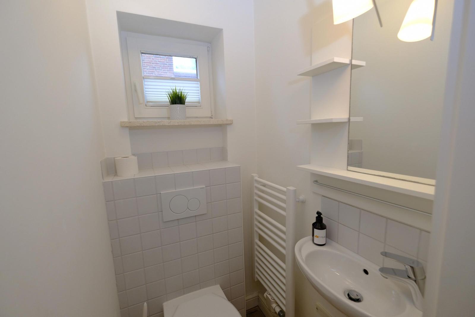 Badezimmer mit Waschbecken, Spiegel, Heizung und Toilette. Fenster mit Pflanze.