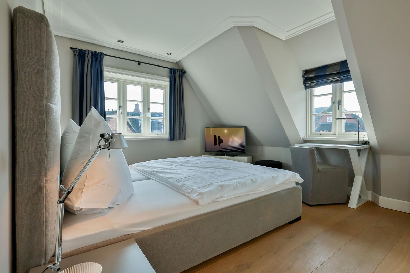 Schlafzimmer mit Bett, Schreibtisch und TV. Zwei Fenster mit Blick nach draußen.