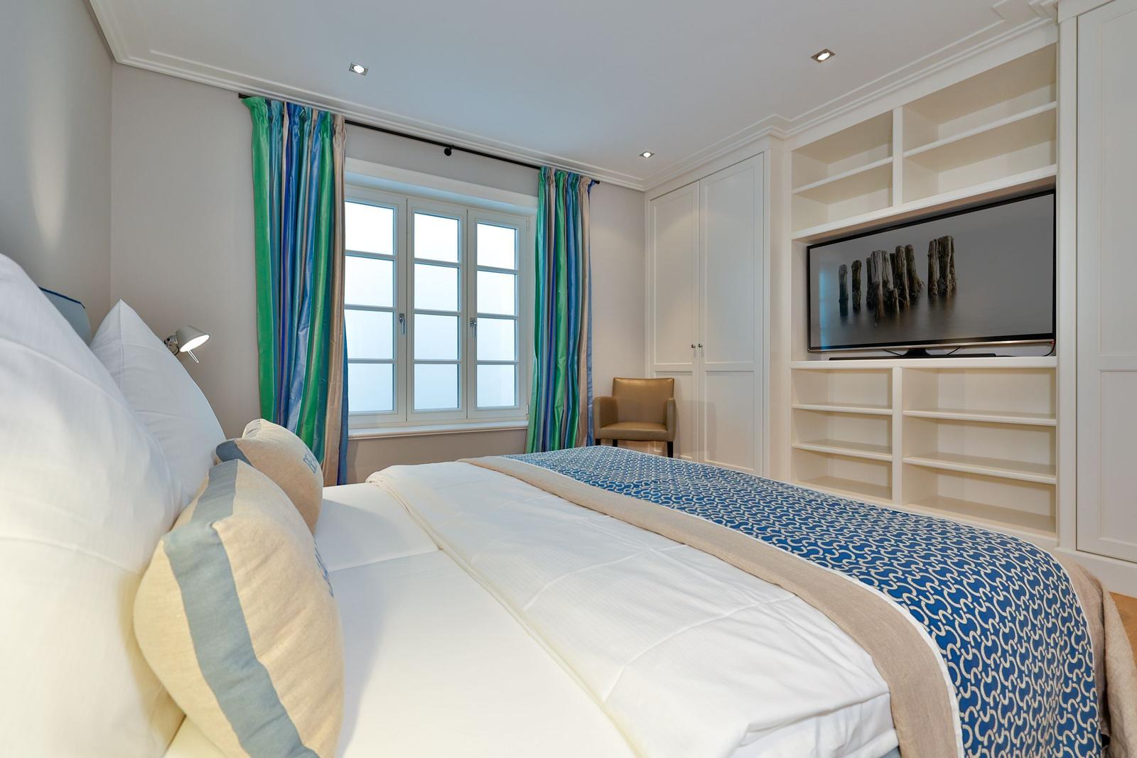 Schlafzimmer mit Bett, Fenster, Gardinen und TV-Wand.