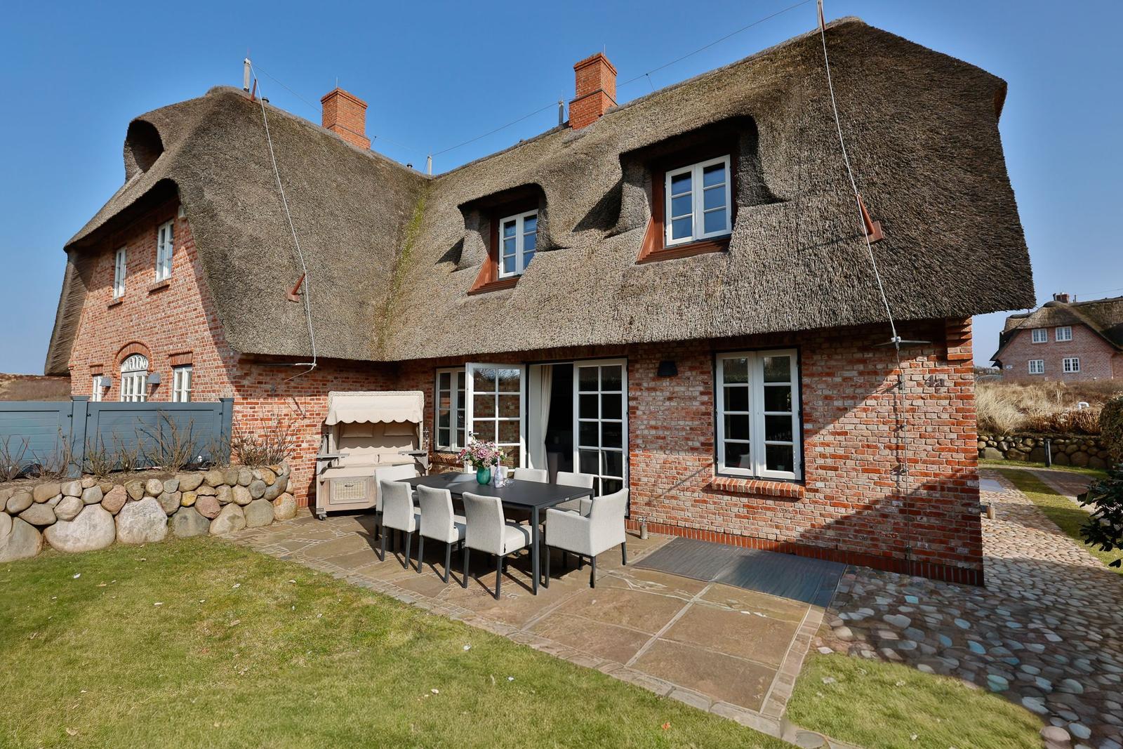 Haus mit Strohdach, Terrasse und Garten. Tisch mit Stühlen und Grill.
