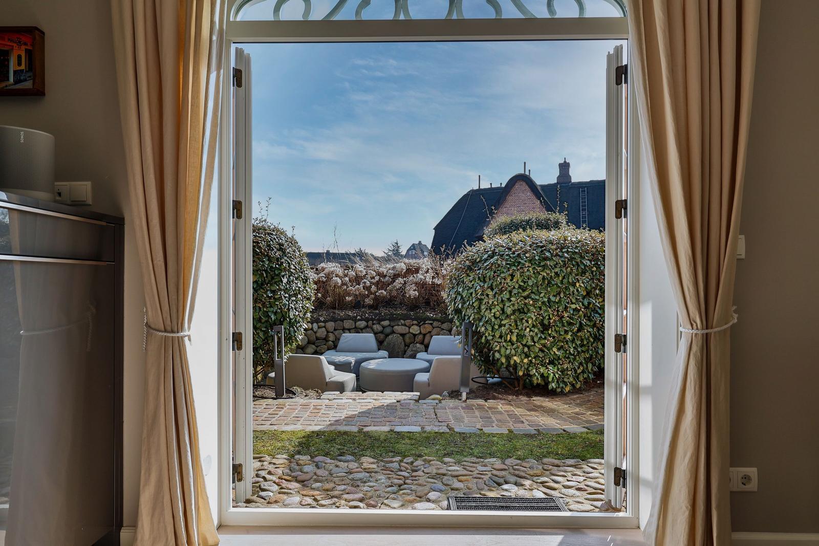Durch die offene Tür blickt man auf eine Terrasse mit Sitzmöbeln und einen gepflasterten Weg.