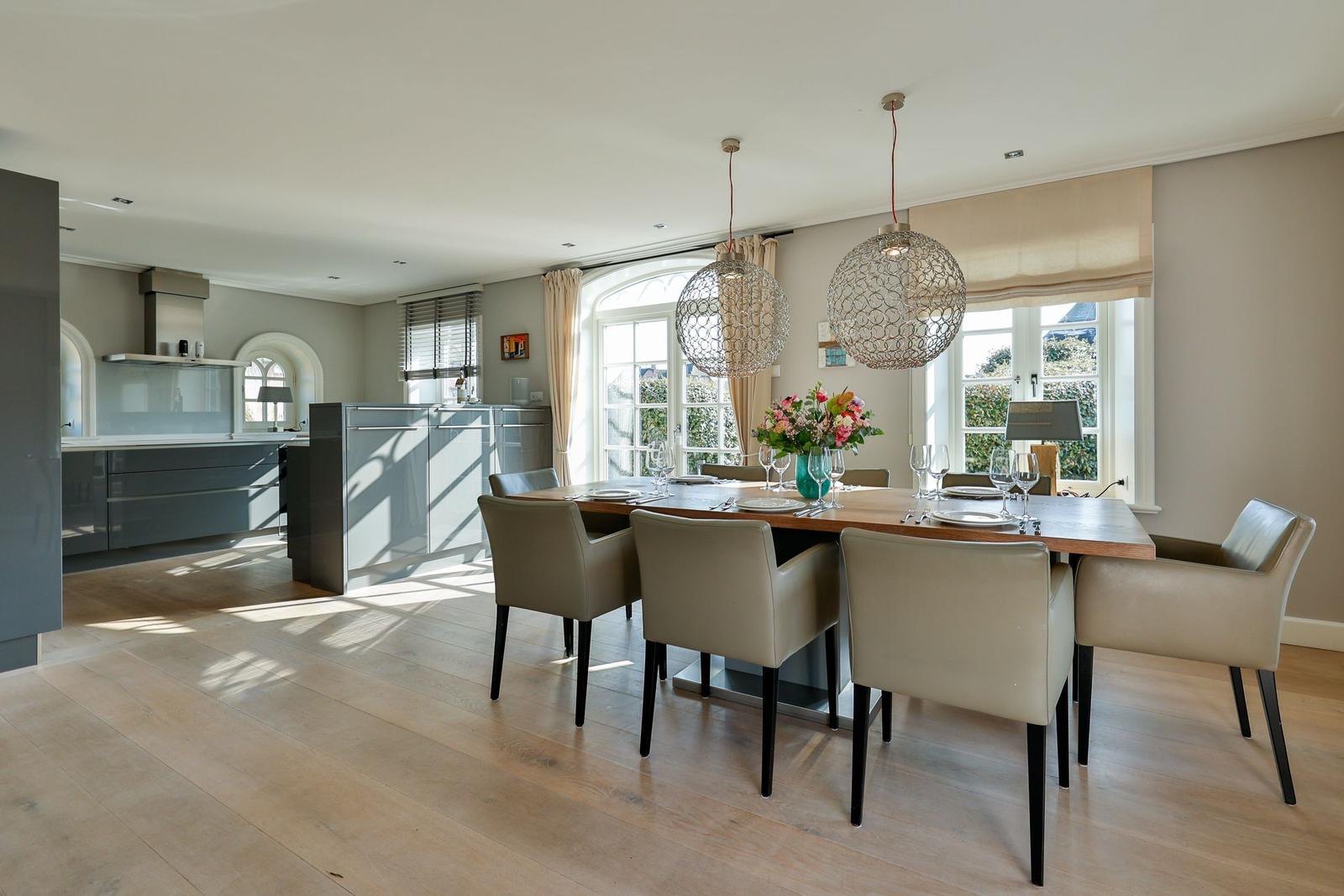 Essbereich mit Tisch, Stühlen und Blumen. Küche im Hintergrund. Holzboden und große Fenster.