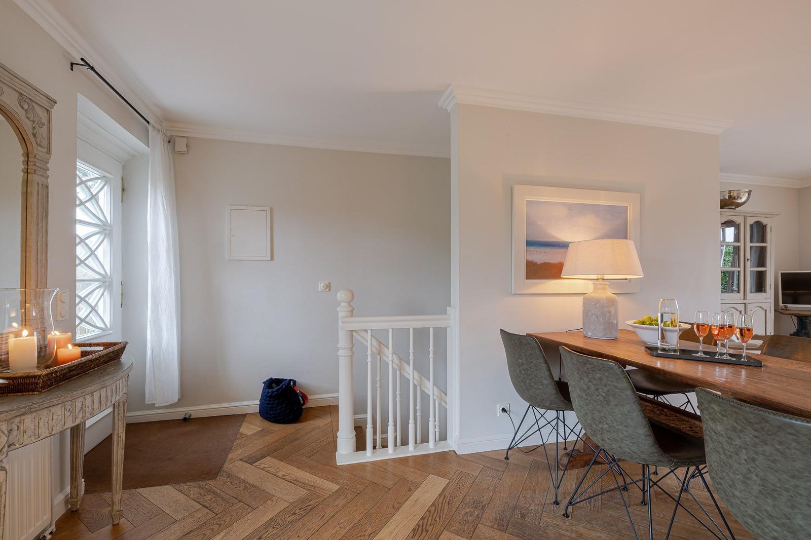 Dining area with wooden table, chairs, and lamp. Light walls, wooden floor, and staircase visible.