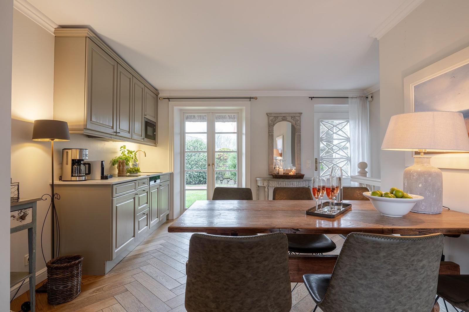 Dining room with wooden table, chairs, and kitchen area. Windows with garden view.