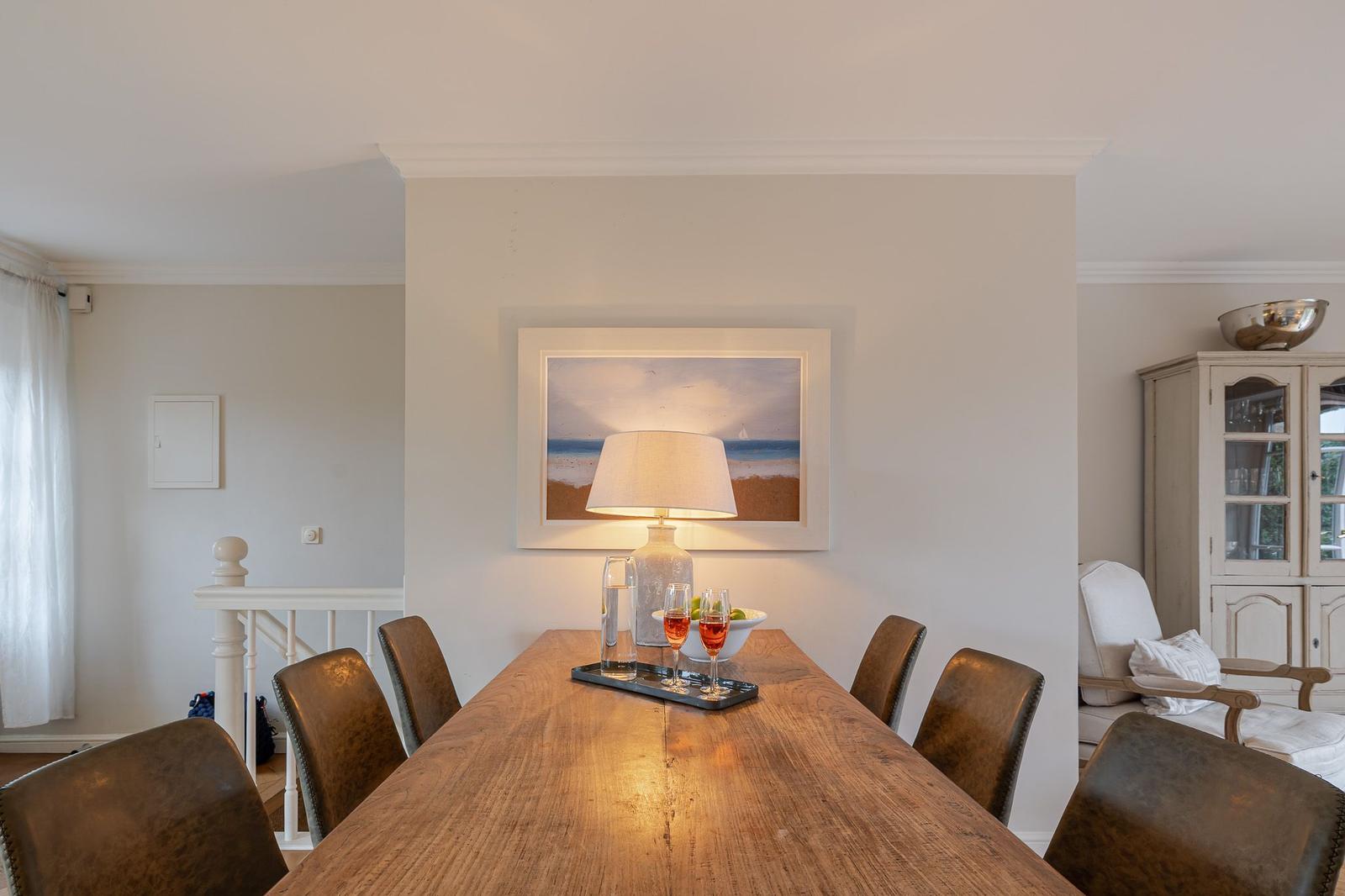 Dining room with wooden table, chairs, and wall art. Lamp and fruit bowl on table.