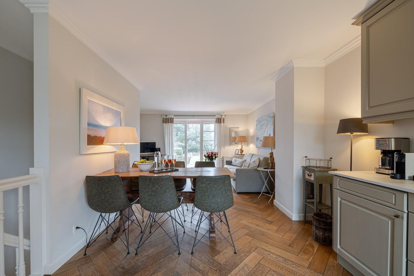 Dining area with table and chairs, living area with sofa and window, kitchen with countertop and coffee machine.