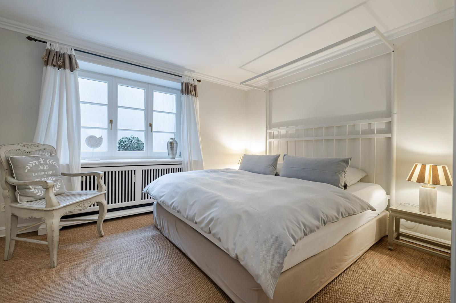 Bedroom with bed, nightstand, and chair. Window with curtains and radiator.