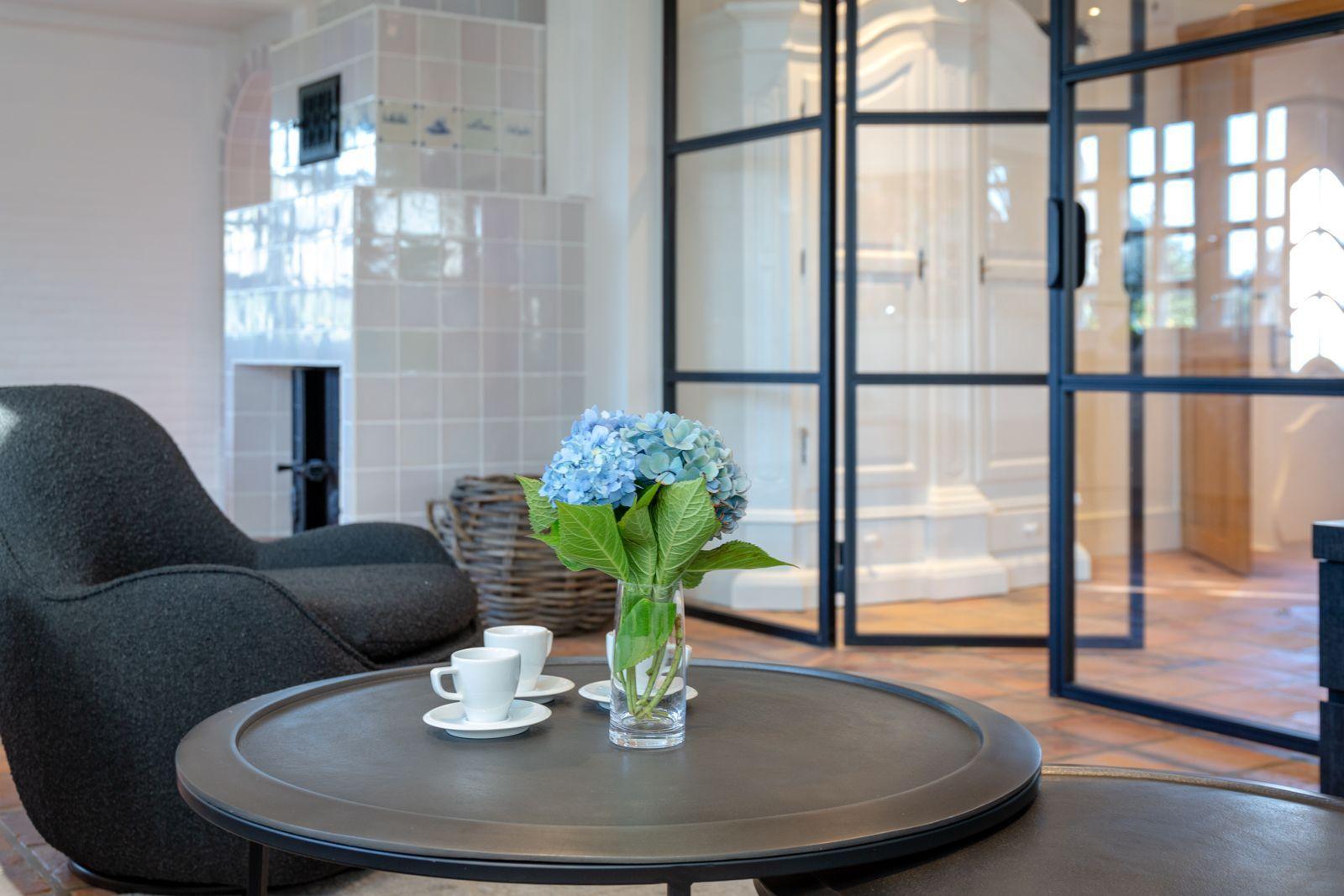 Round table with flowers and cups in front of a chair and fireplace.