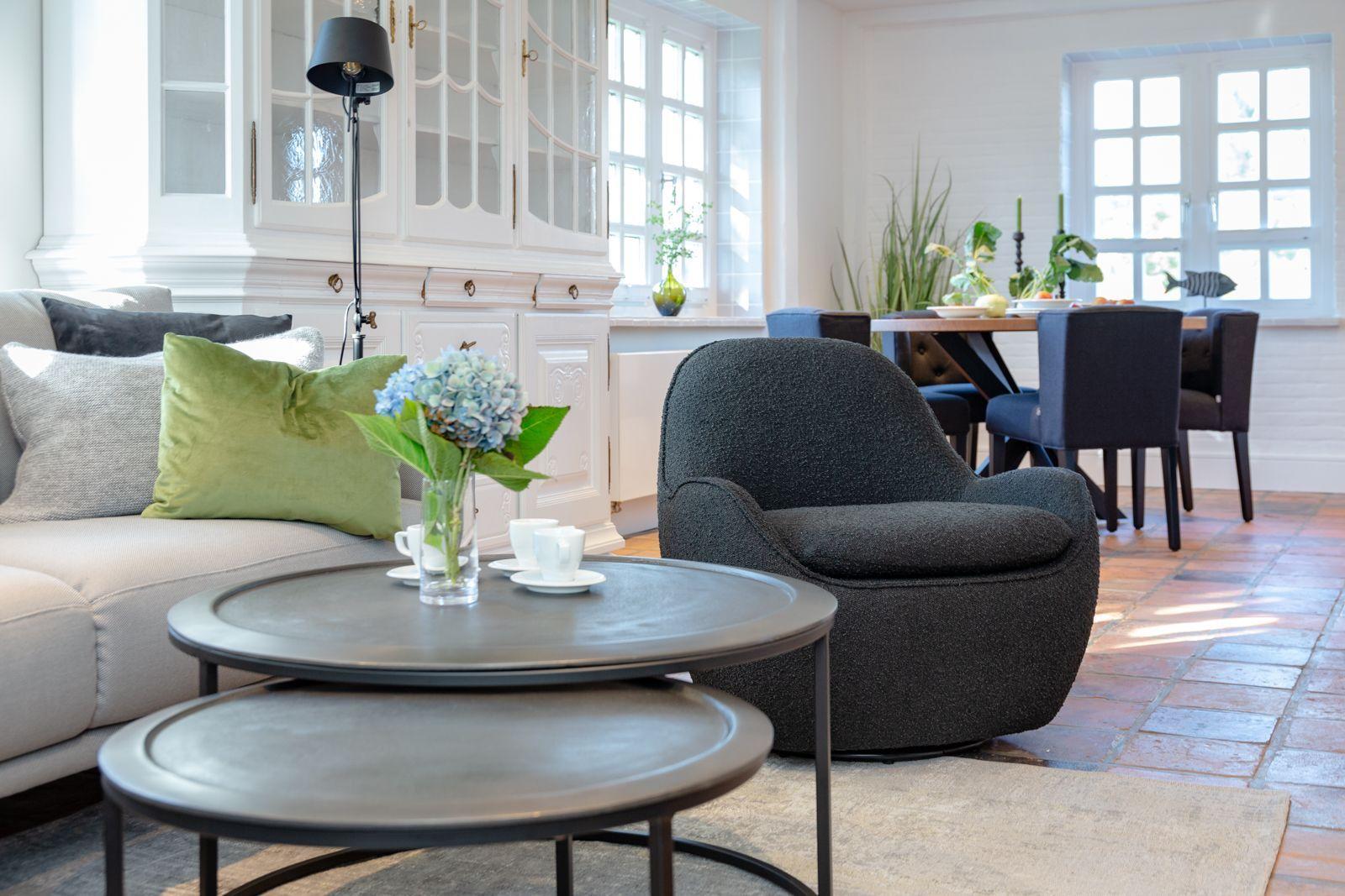 Living room with sofa, armchair, and dining table. Wooden floor and windows visible.