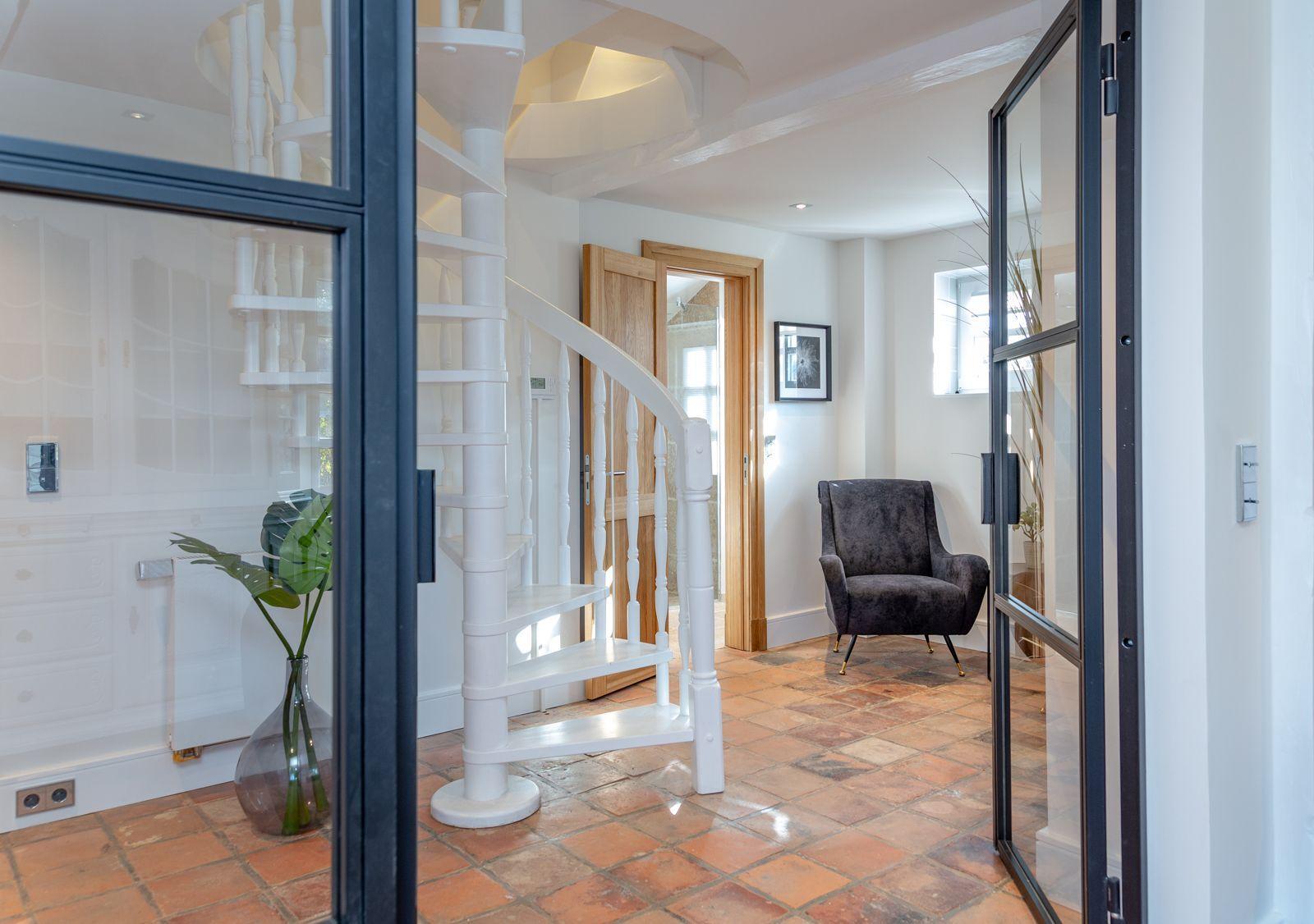 Entrance with white spiral staircase and terracotta floor. Dark armchair and glass walls.