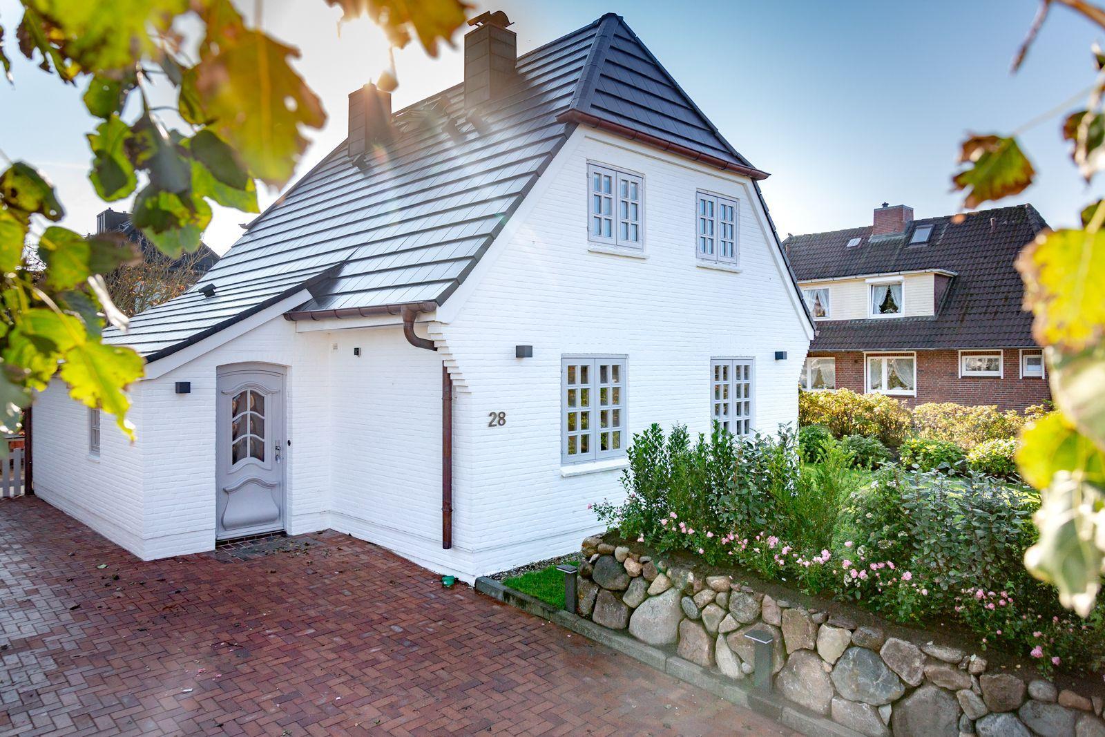 White villa with gray roof and paved driveway. Garden with stone wall and flowers.