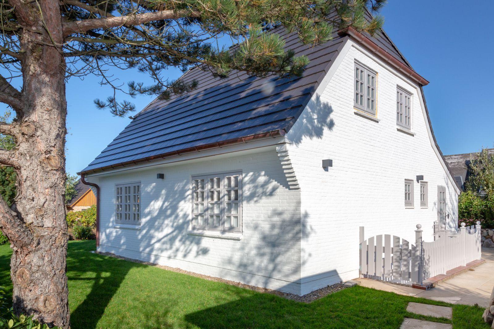 White villa with gray windows and dark roof. Green lawn and tree in front of the house.