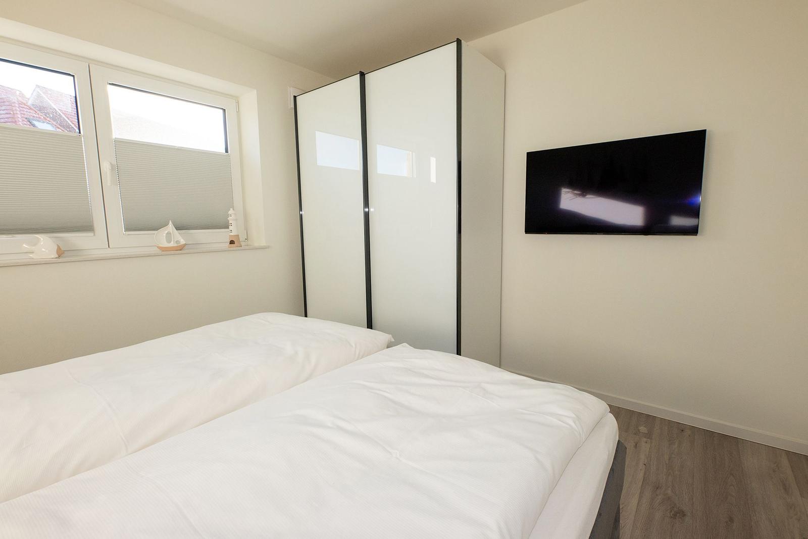 Schlafzimmer mit Doppelbett, Kleiderschrank und Wandfernseher. Fenster mit Jalousie.