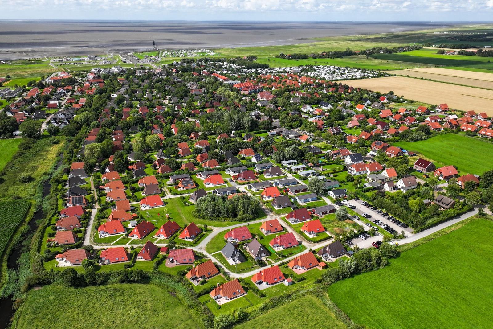 Luftbild eines Ortes mit Häusern, Grünflächen und einem Strand im Hintergrund.