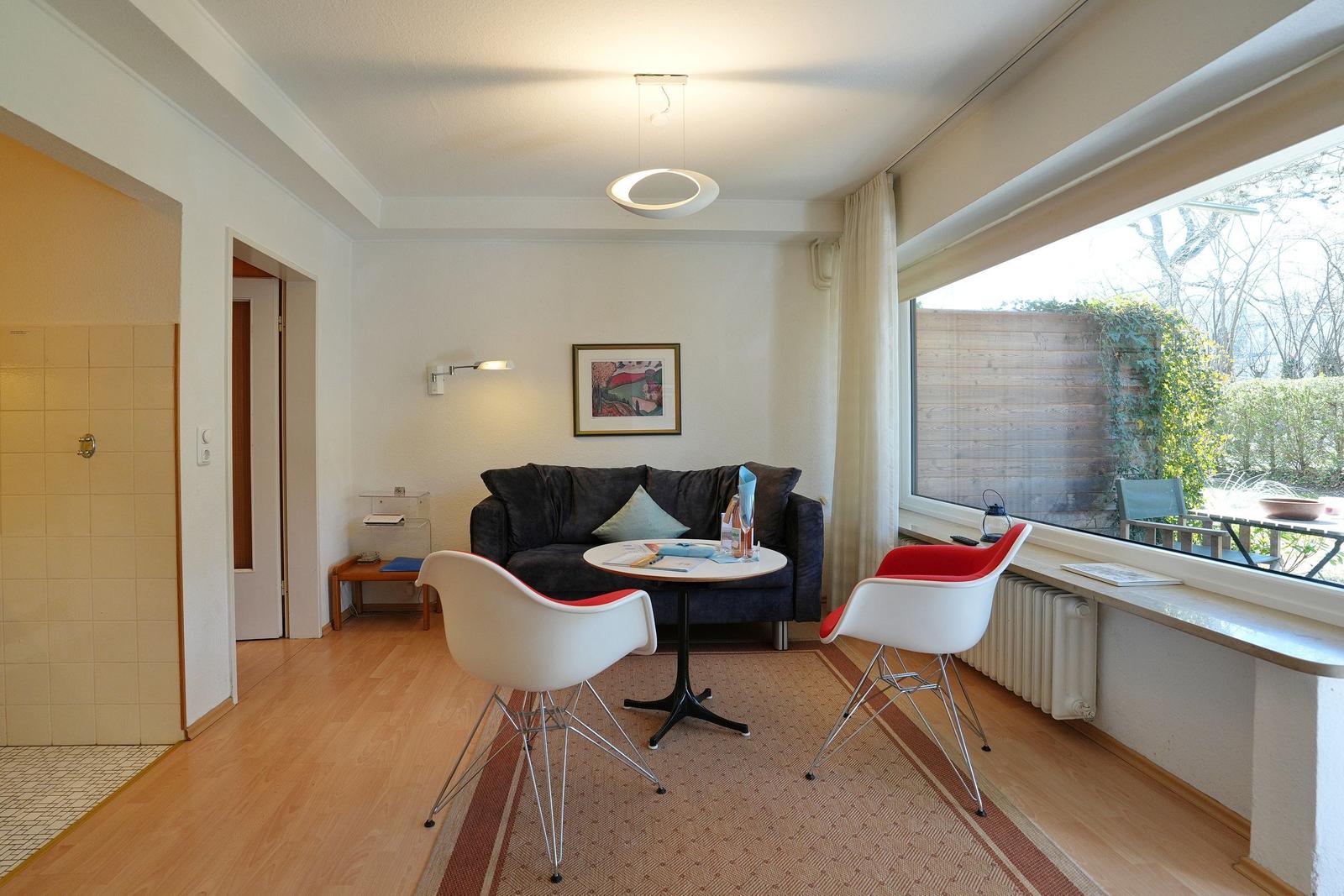 Wohnzimmer mit Sofa, zwei Stühlen und großem Fenster. Blick auf Garten. Flur und Bad sind sichtbar.