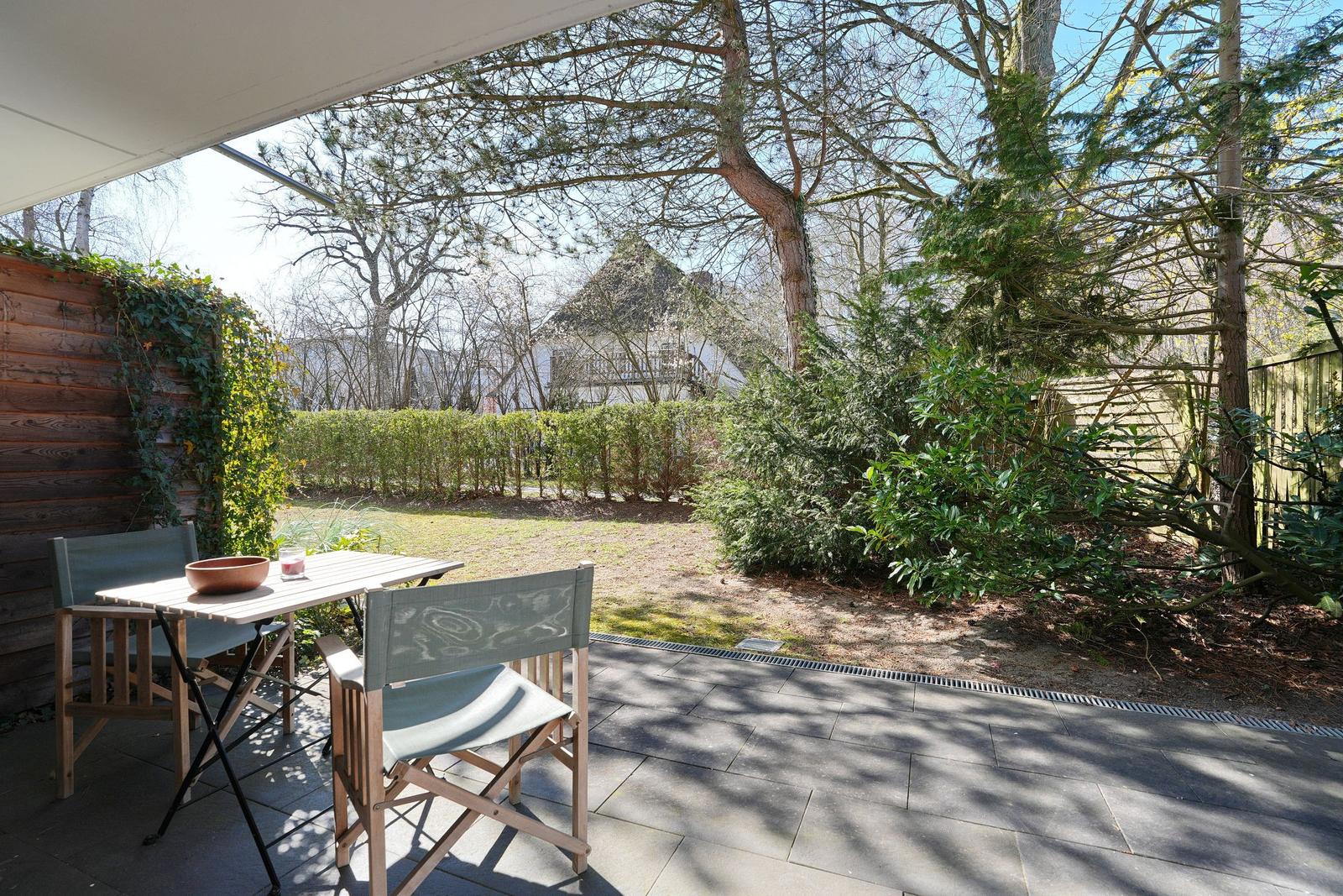 Terrasse mit Tisch und Stühlen, Blick auf Garten mit Bäumen und Hecke.