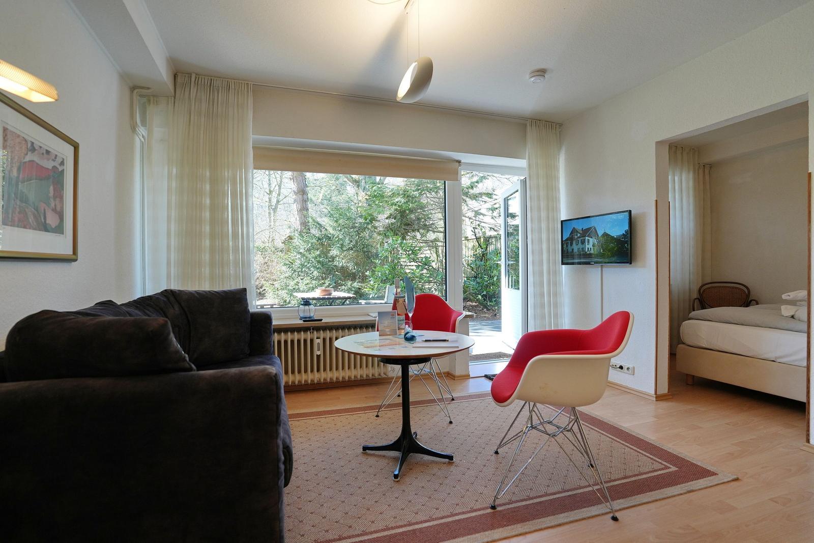 Wohnraum mit Sofa, Tisch, zwei Stühlen und Schlafzimmer im Hintergrund. Fenster mit Blick auf Garten.