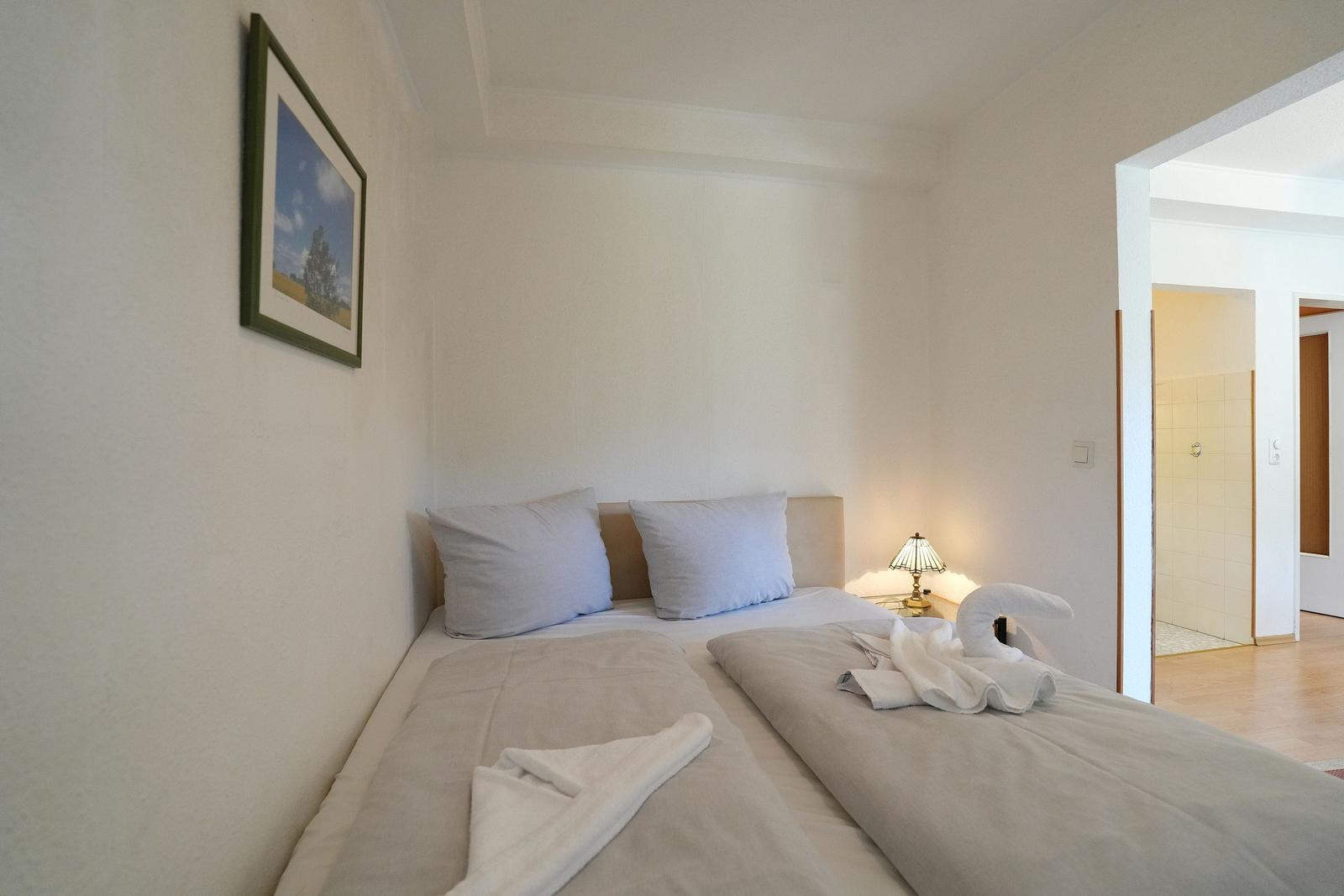 Schlafzimmer mit Bett, Kissen, Nachttisch und Lampe. Ein Schwanz aus Handtuch liegt auf dem Bett.