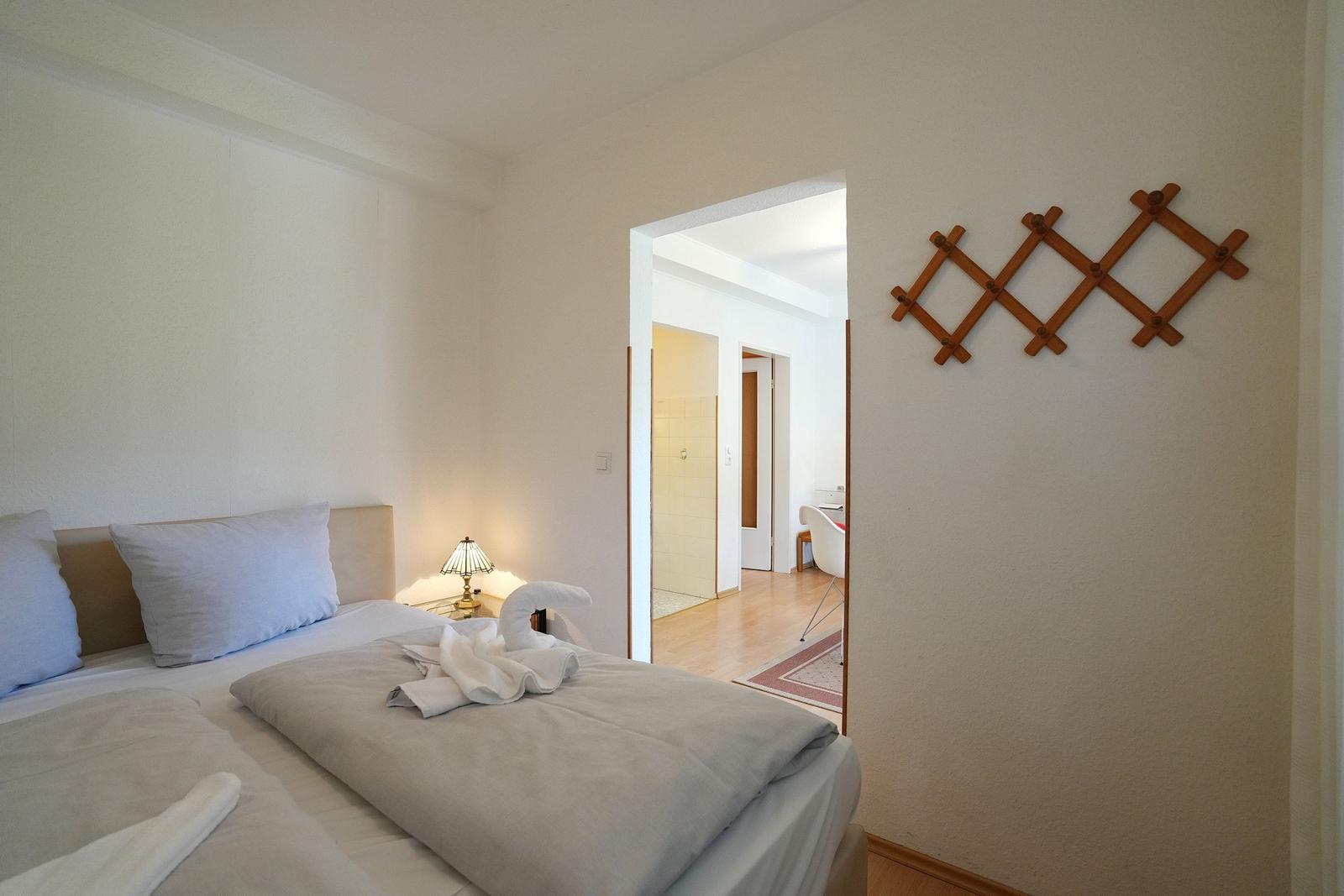 Schlafzimmer mit Bett, Nachttisch und Lampe. Durchgang zu Bad und Wohnbereich. Wanddekoration aus Holz.
