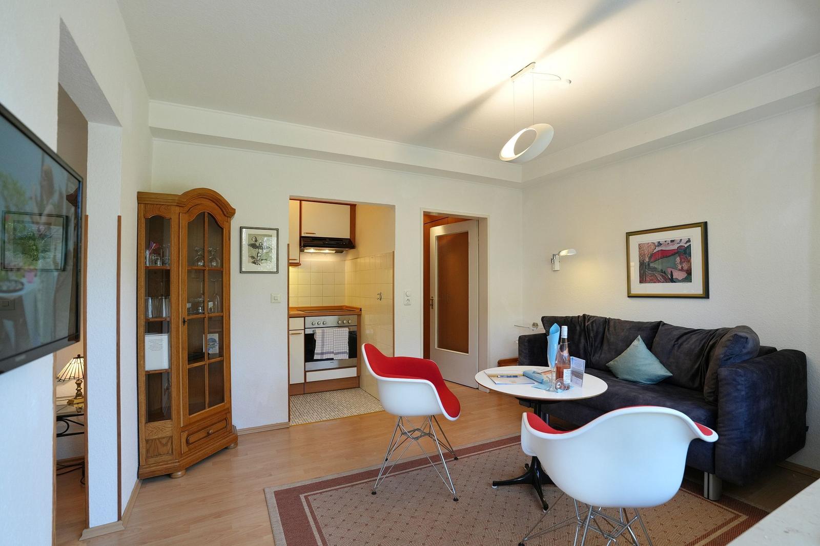 Wohnzimmer mit Sofa, Tisch und zwei Stühlen. Küche im Hintergrund. Holzfußboden und Wandbild.