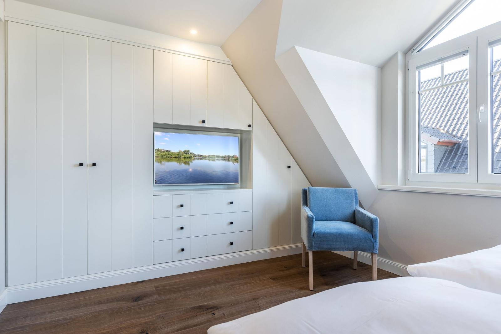Bedroom with blue armchair, TV, and window. Wooden floor and white cabinets.