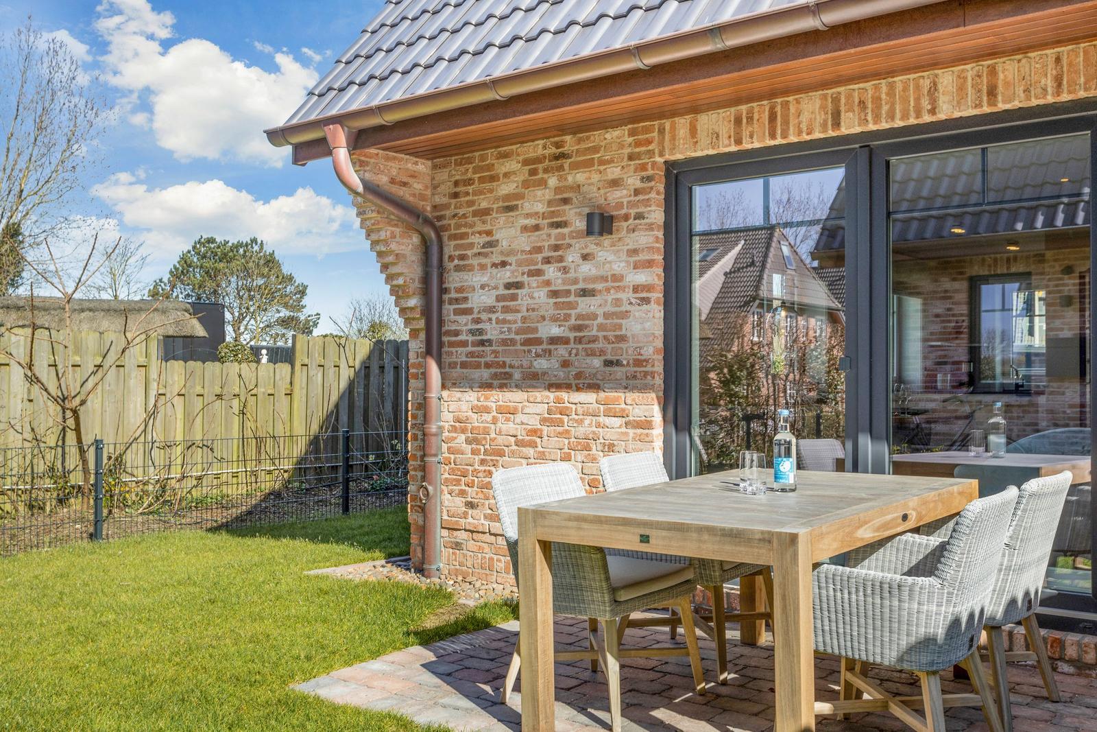 Terrace with wooden table and chairs beside brick house with garden gate.