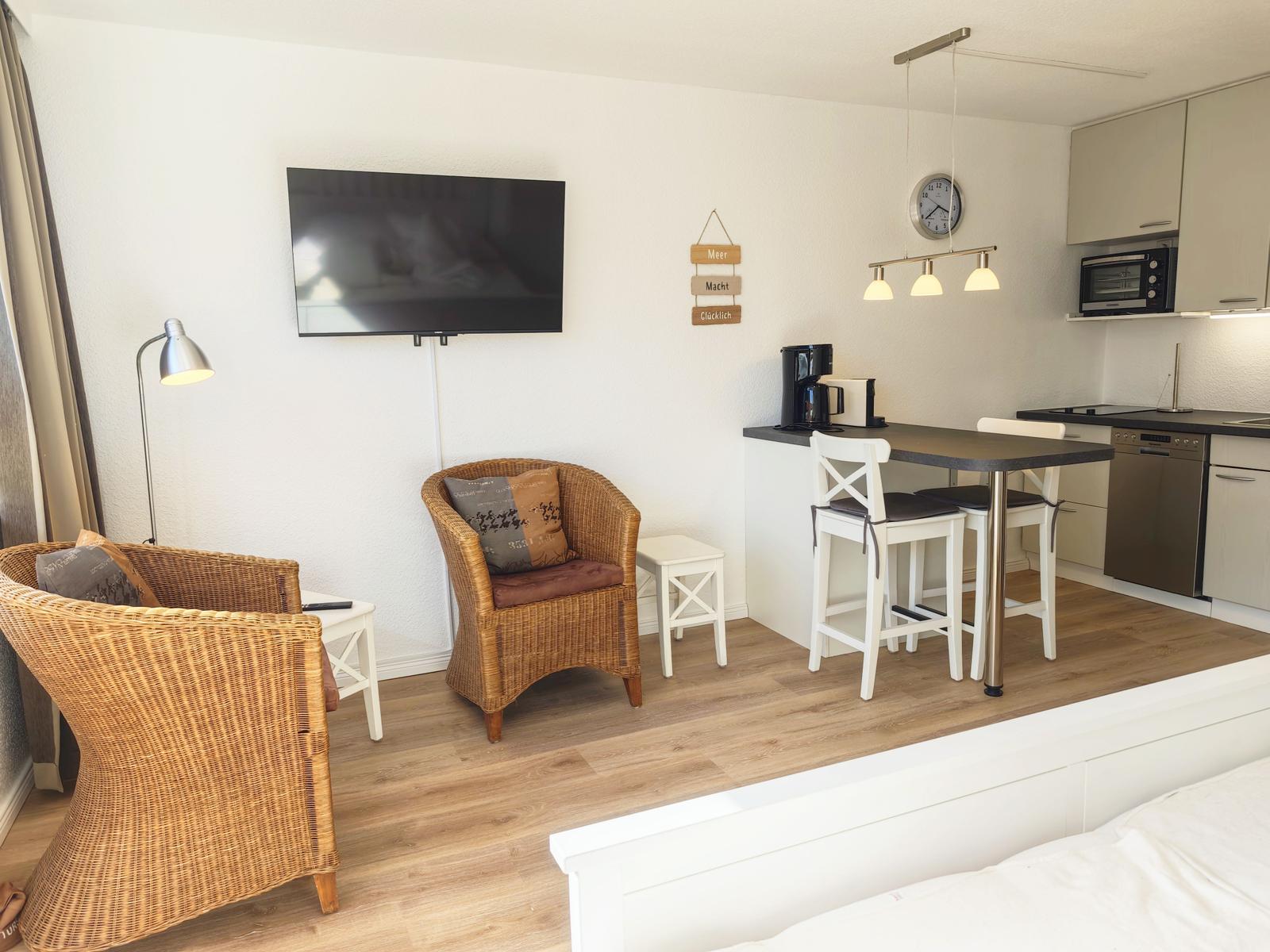 Wohnbereich mit TV, Esstisch und Küche. Holzdielenboden und Wickerstühle.